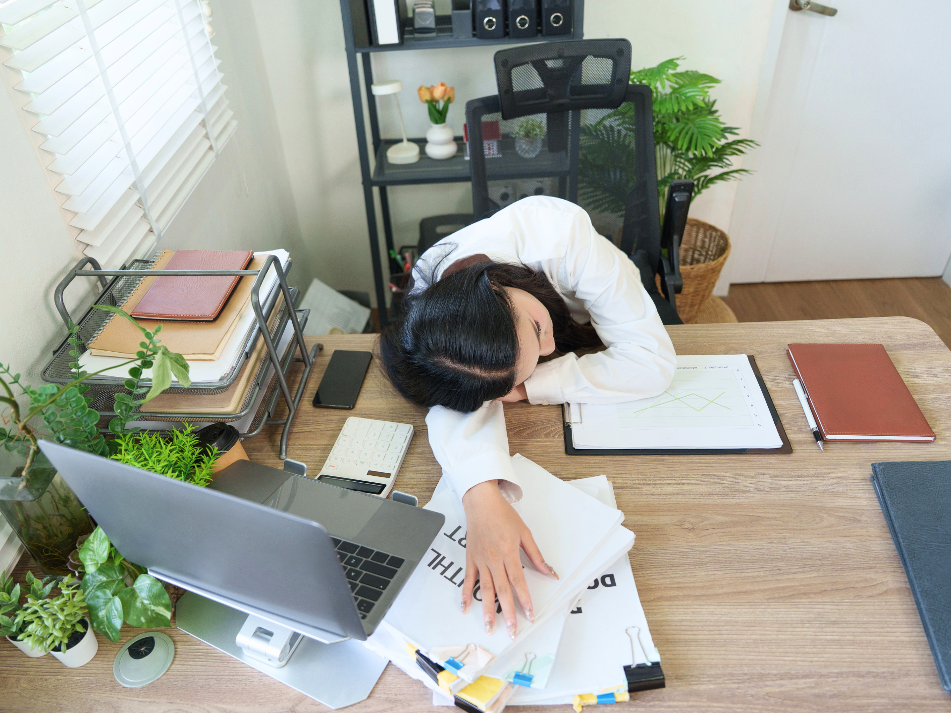 Person slumped over a desk in a bright office, surrounded by laptop, papers, and plants