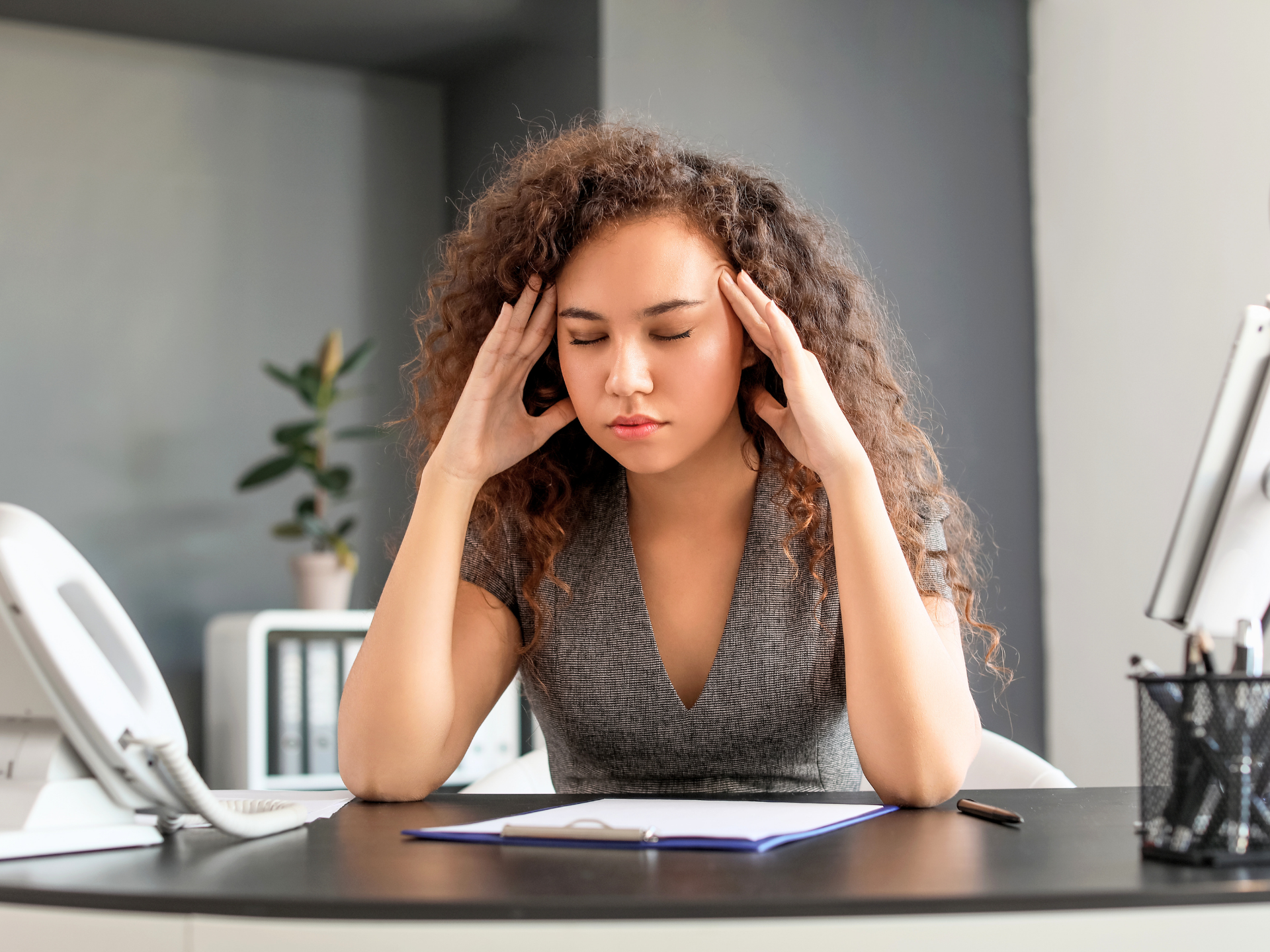 Woman holding her temples at a desk with papers, looking stressed in an office