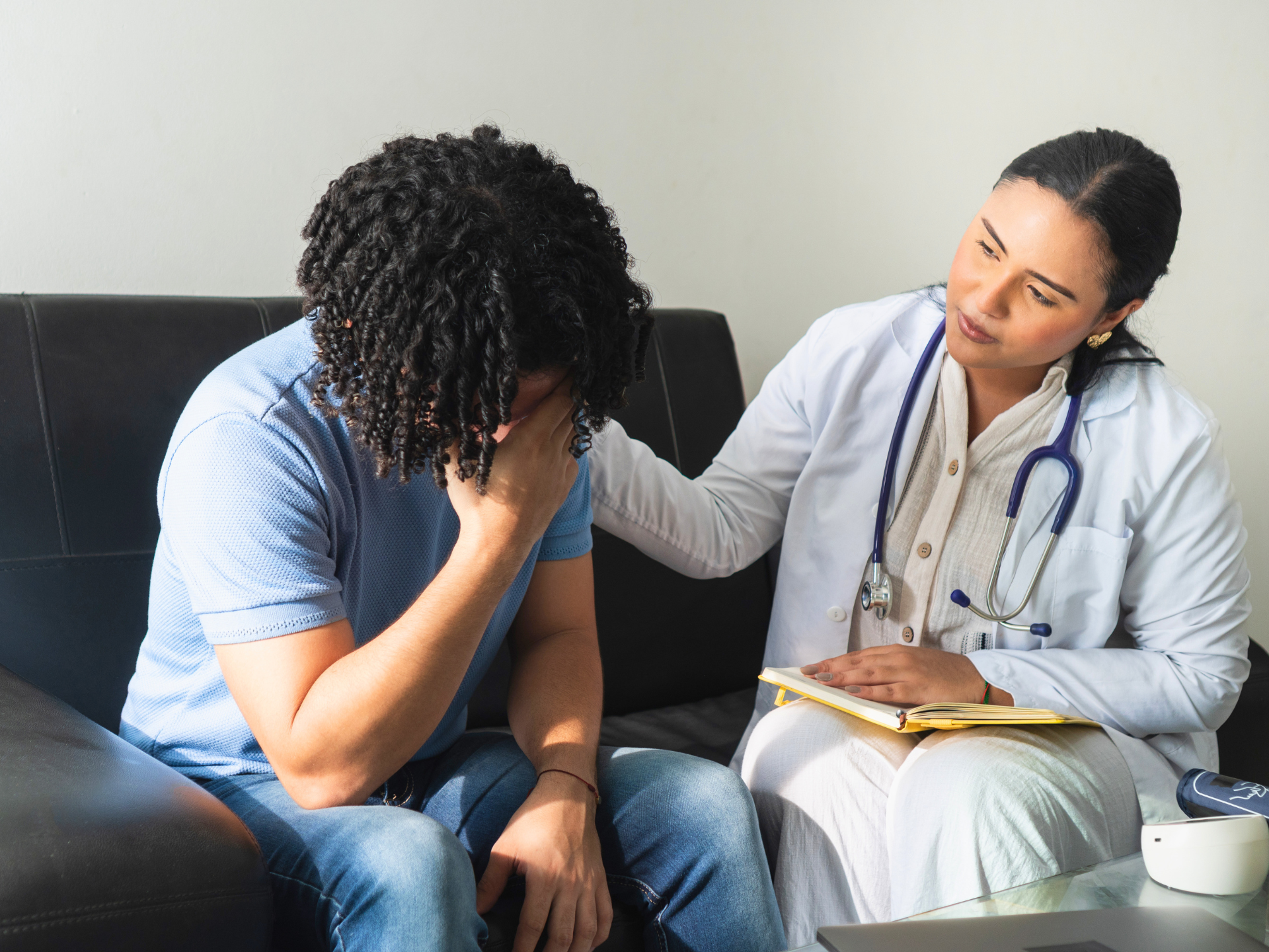 Patient seated on couch, covering face, while clinician in white coat offers support in a counseling session