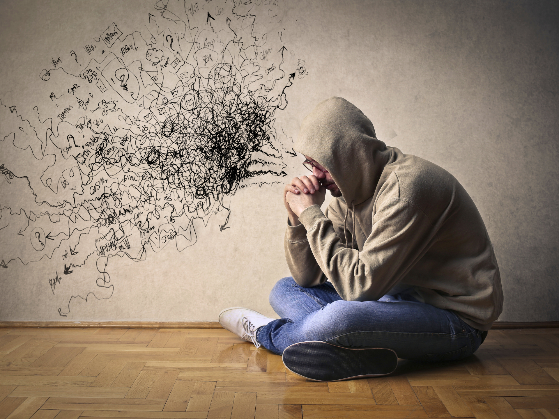 Hooded person sits on the floor with head in hands as black scribbles burst from the wall nearby