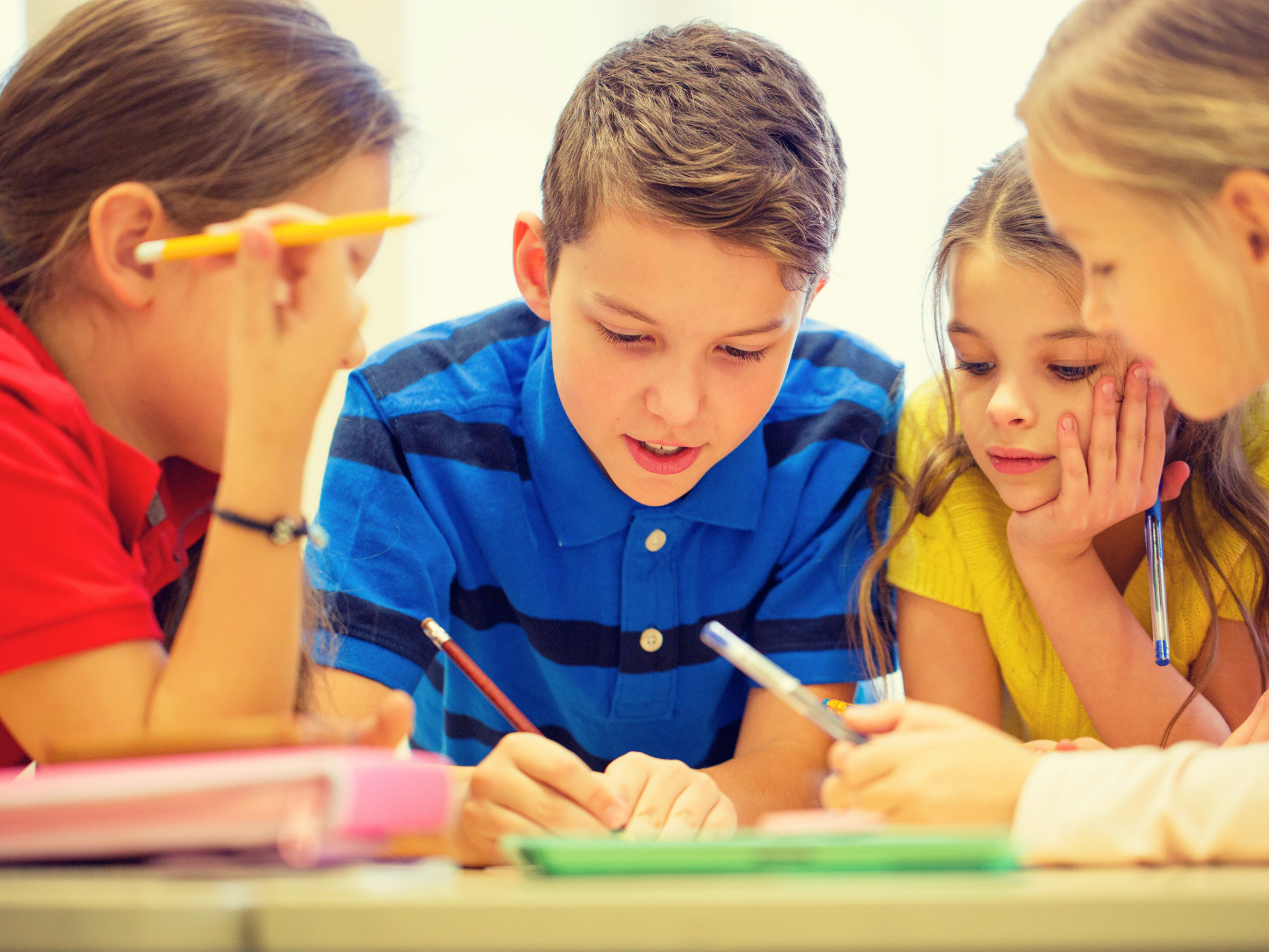 Three children lean over a table, reading and writing together in a bright classroom.