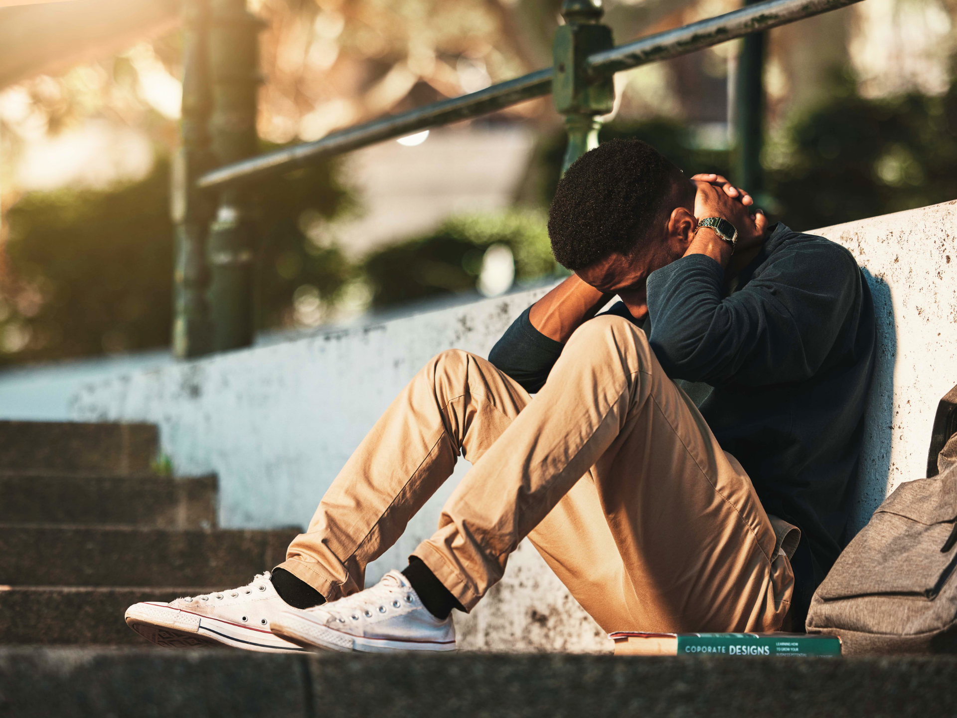 Person sitting on outdoor steps, knees hugged to chest, head resting on arms, in warm sunlight.