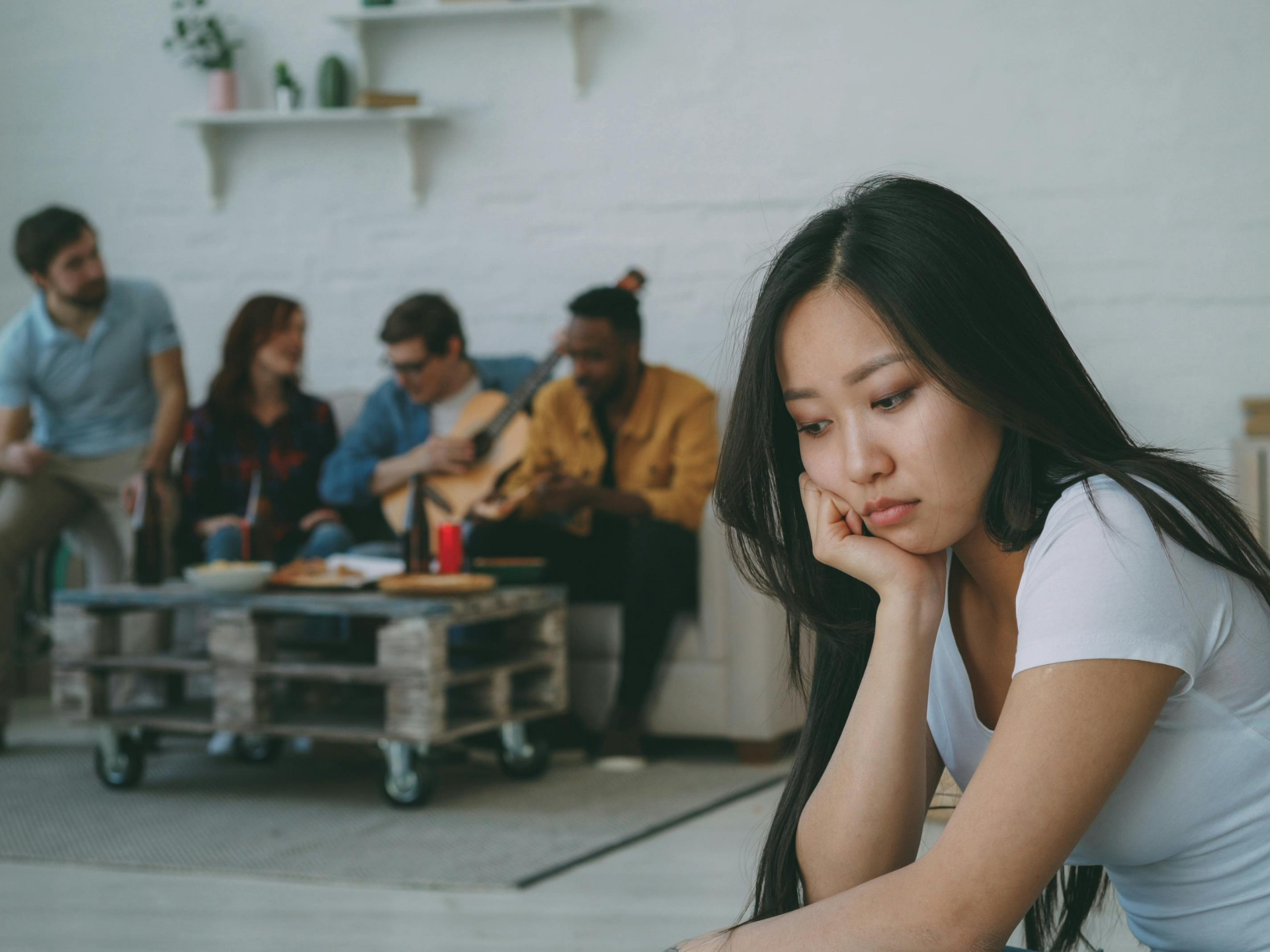 Woman sitting apart and looking down while a group chats on a couch in the background