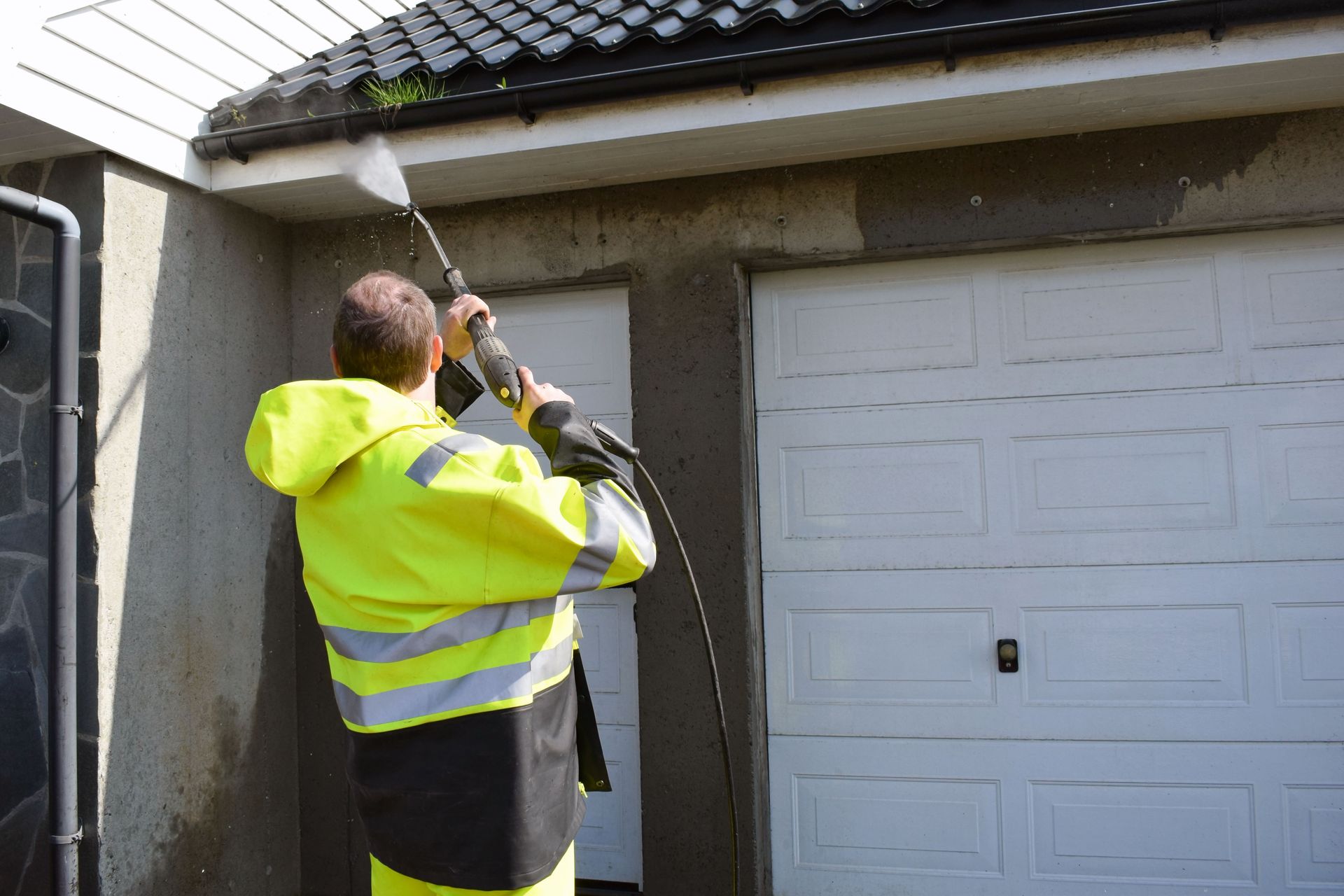 A man in a yellow jacket is using a high pressure washer to clean a garage door.