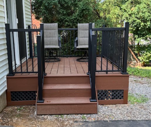Small front porch with black railings, two chairs, and brown steps beside a house.