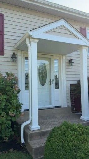 Front porch entrance with white columns and door on a beige house, bordered by shrubs.