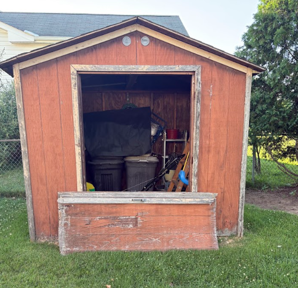 Open wooden shed with a large barrel and tools inside, front door propped open in a grassy yard.