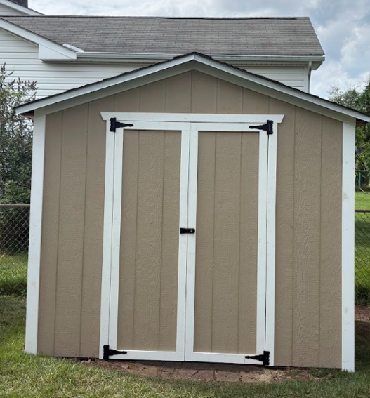 Tan shed with white double doors and black hinges in a backyard beside a house.