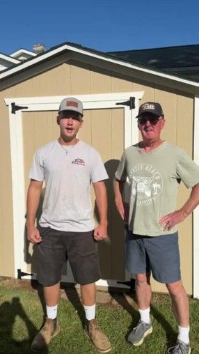 Two men standing in a yard in front of a tan shed, wearing casual clothes and hats.