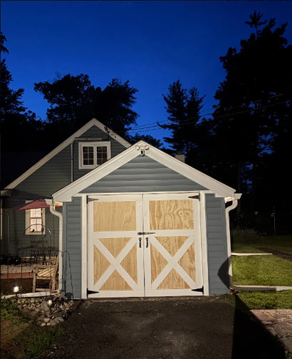 Gray garage with white X doors and a dark driveway at dusk, framed by trees and a deep blue sky.