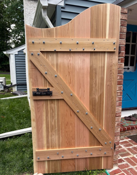 Wooden garden gate with diagonal brace and curved top, set beside a brick house and blue door.
