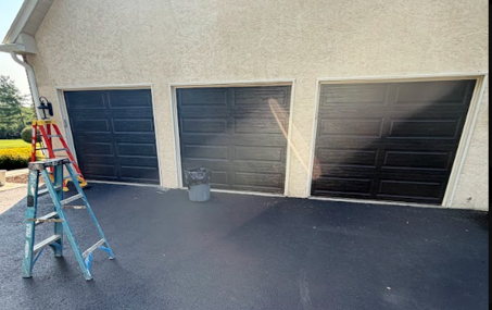 Three black garage doors with a blue ladder and paint bucket on the driveway, sunlight streaking across the wall.