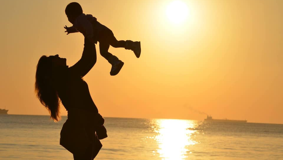 A woman is holding a baby in the air on the beach at sunset.
