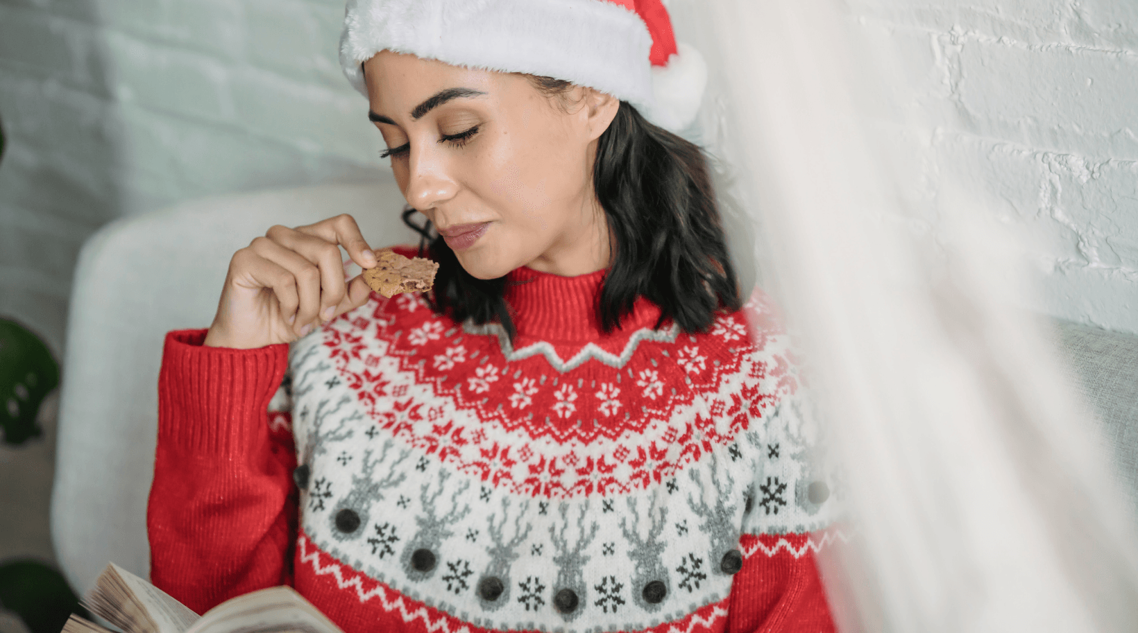 A woman in a santa hat is eating a cookie while reading a book.