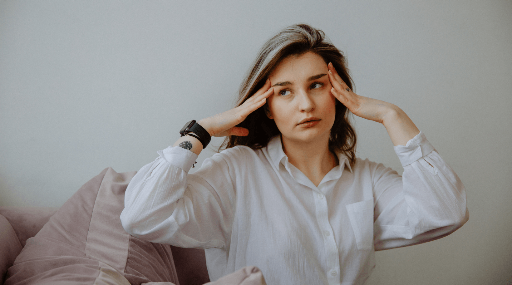 A woman is sitting on a couch with her hands on her head.