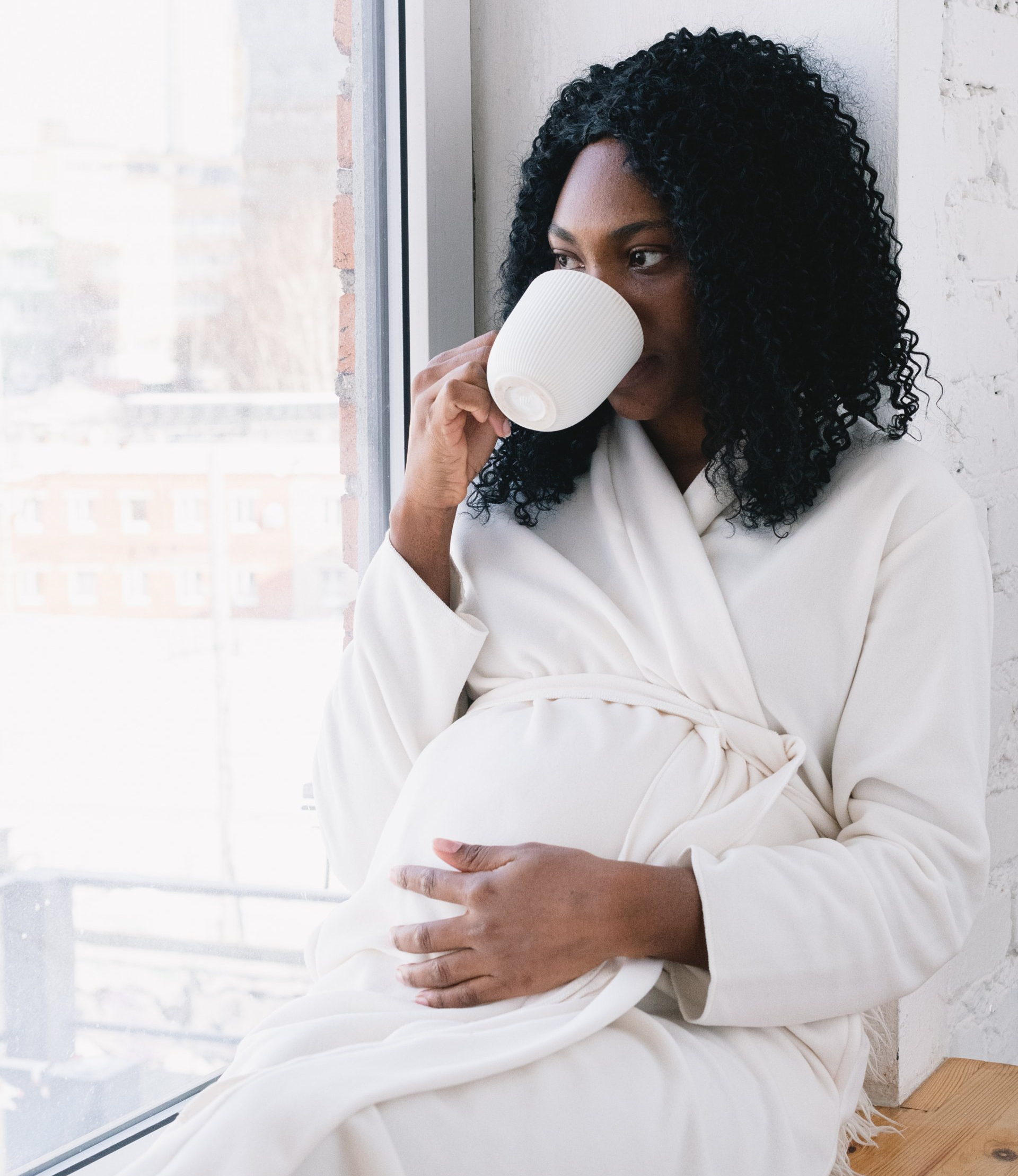 A pregnant woman is drinking a cup of coffee while sitting on a window sill.