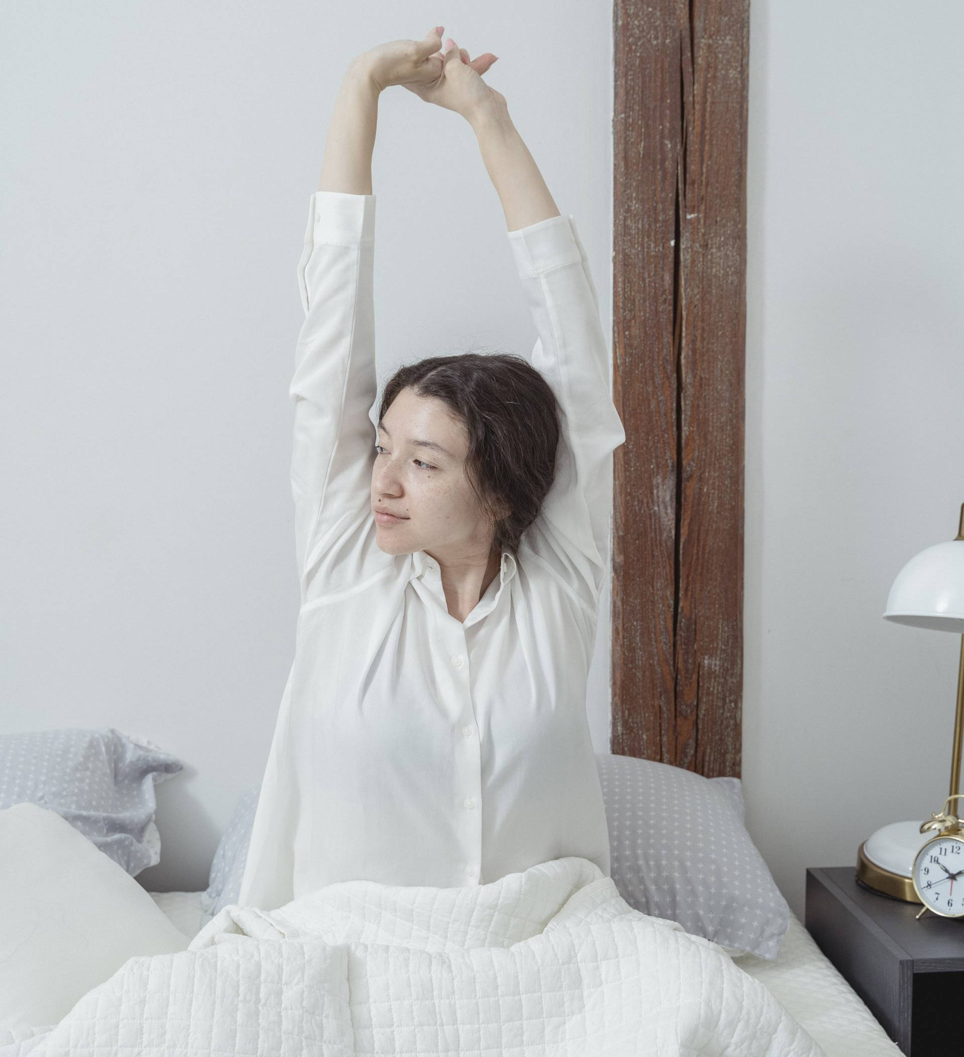 A woman in a white shirt is stretching her arms in bed.