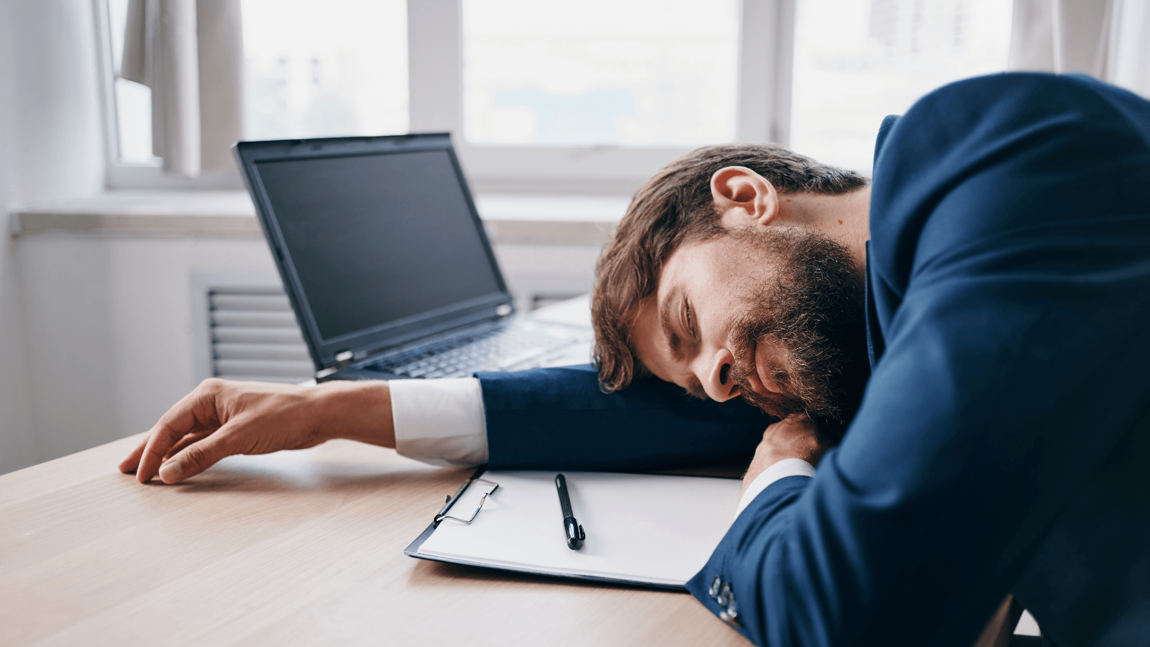 A man is sleeping at a desk in front of a laptop computer.