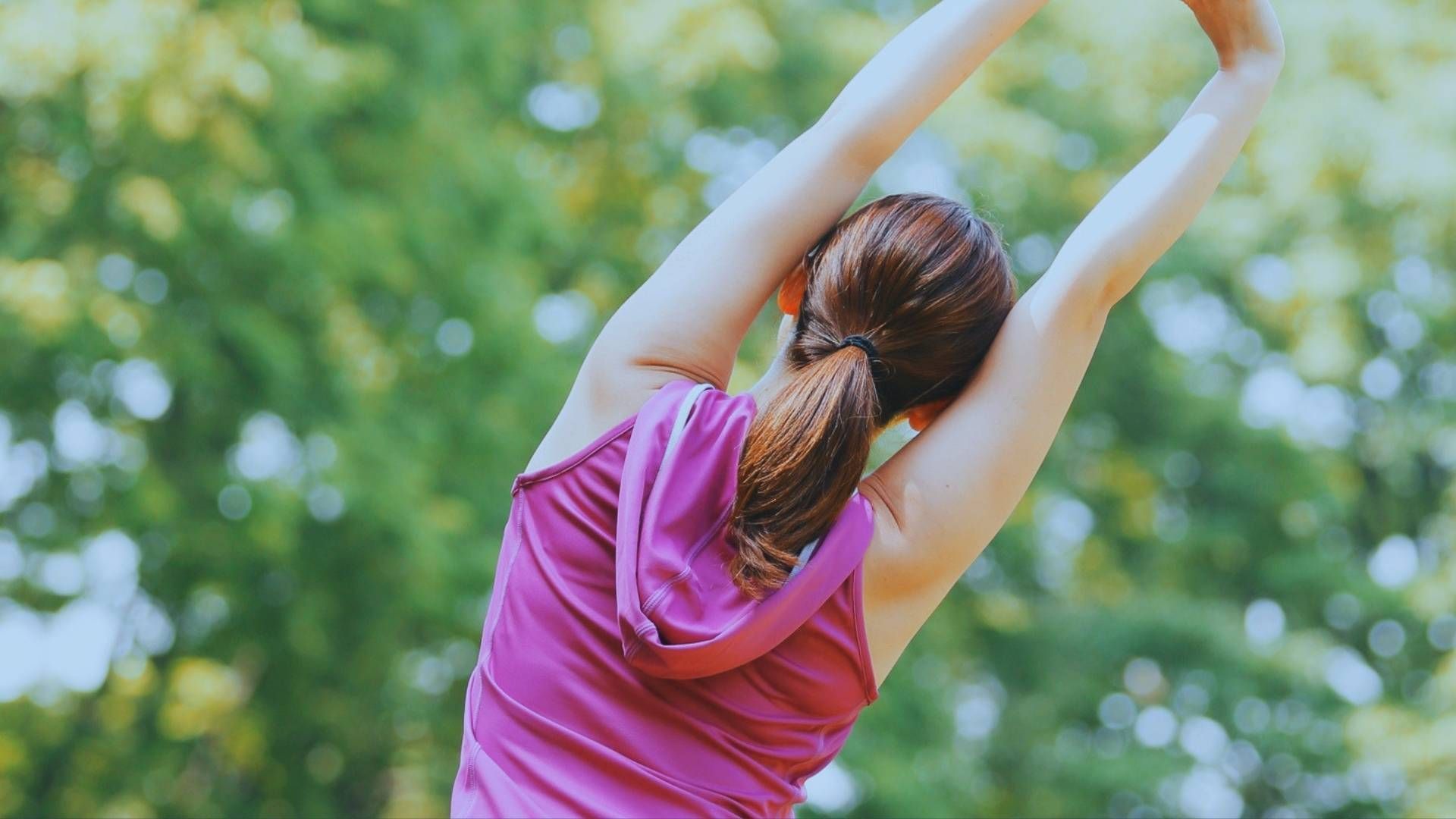 Person stretching outdoors with arms raised overhead, surrounded by soft green summer foliage.