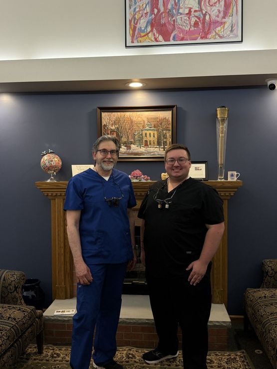 Two people standing smiling in front of a fireplace in a blue-walled room with artwork and decor. Two people standing smiling in front of a fireplace in a blue-walled room with artwork and decor.