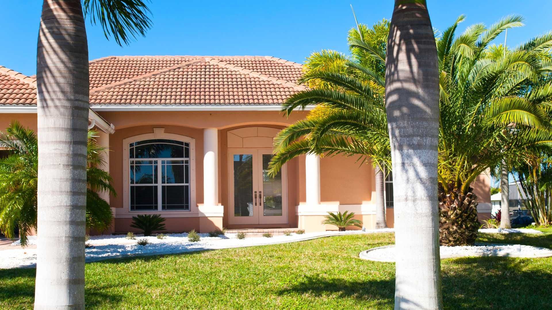 Luxury stucco villa with balconies and lush landscaping, framed by palm trees under a bright blue sky