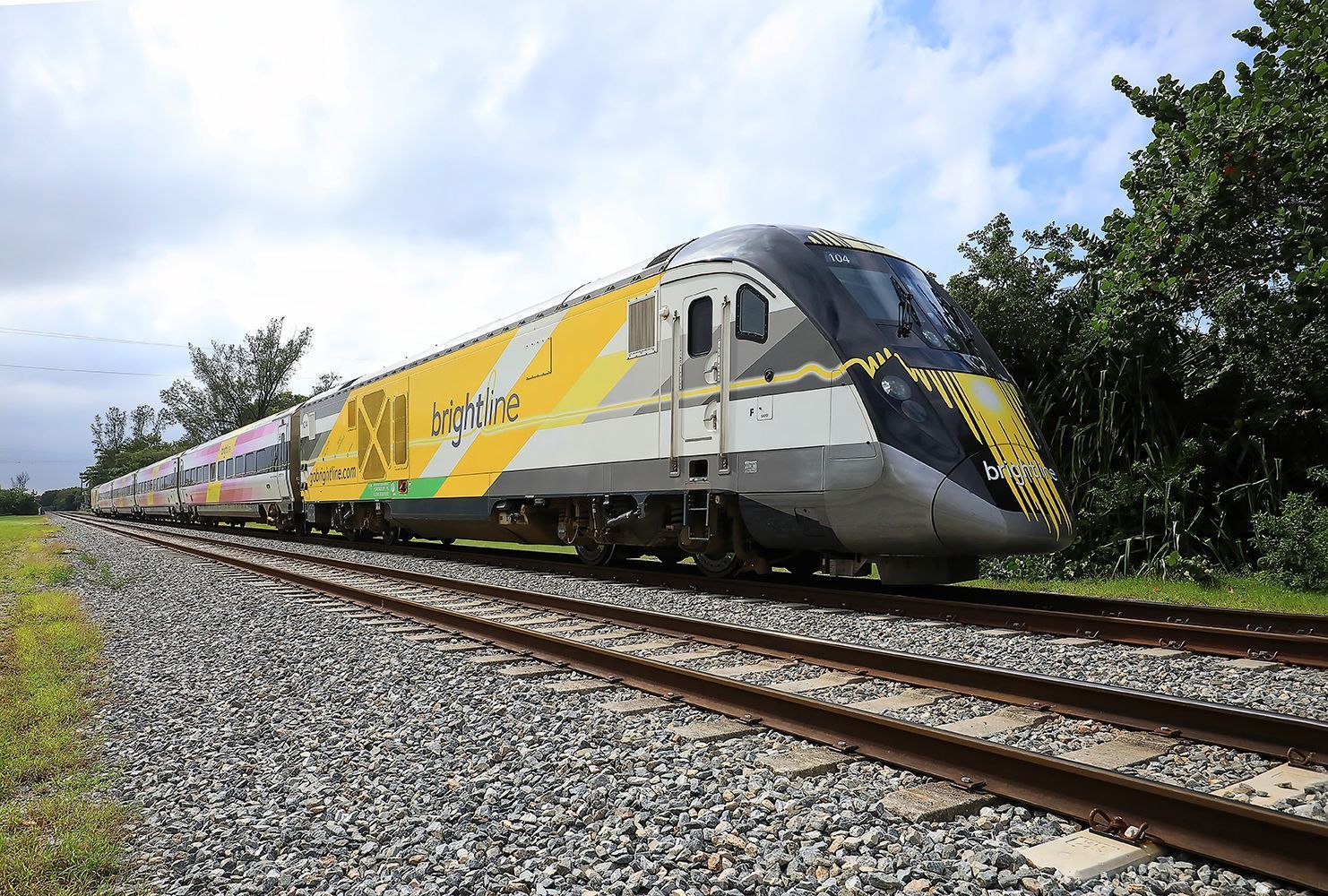 Yellow-and-silver train speeding along tracks beside trees under a blue sky