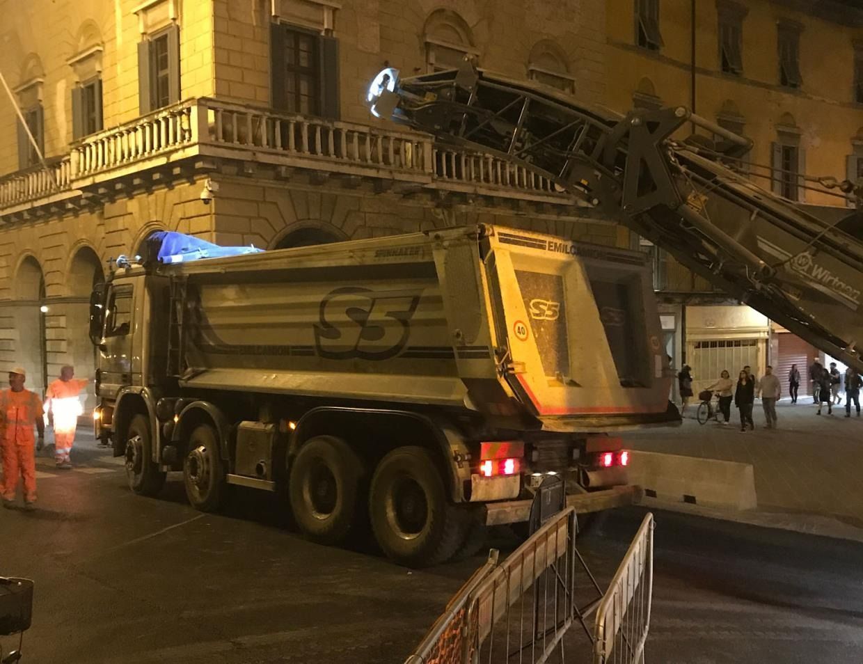 Un autocarro con cassone ribaltabile caricato con asfalto da una fresatrice stradale di notte.
