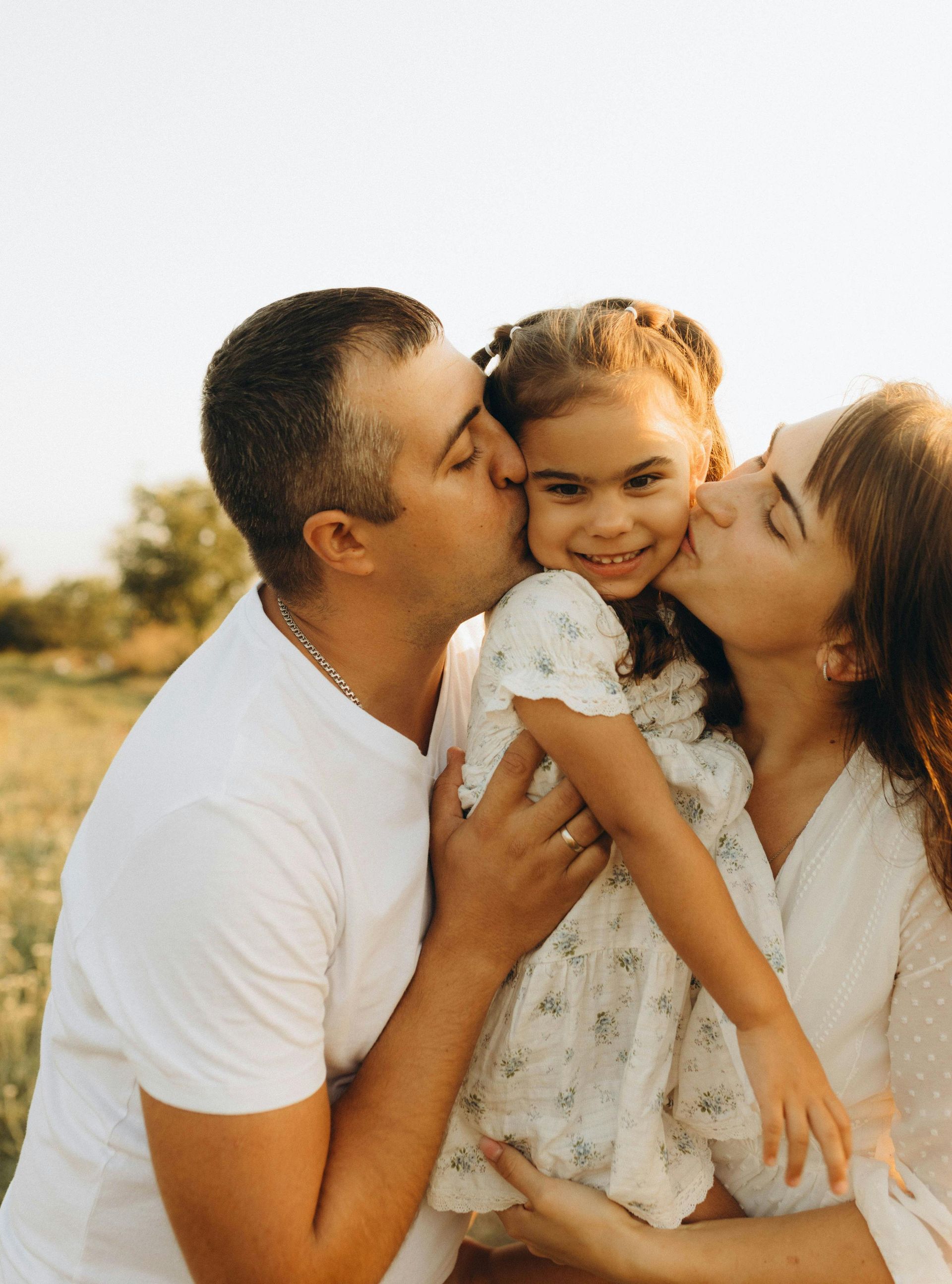 Two parents kiss their smiling young child on the cheeks outdoors in a grassy field, both wearing white clothing.