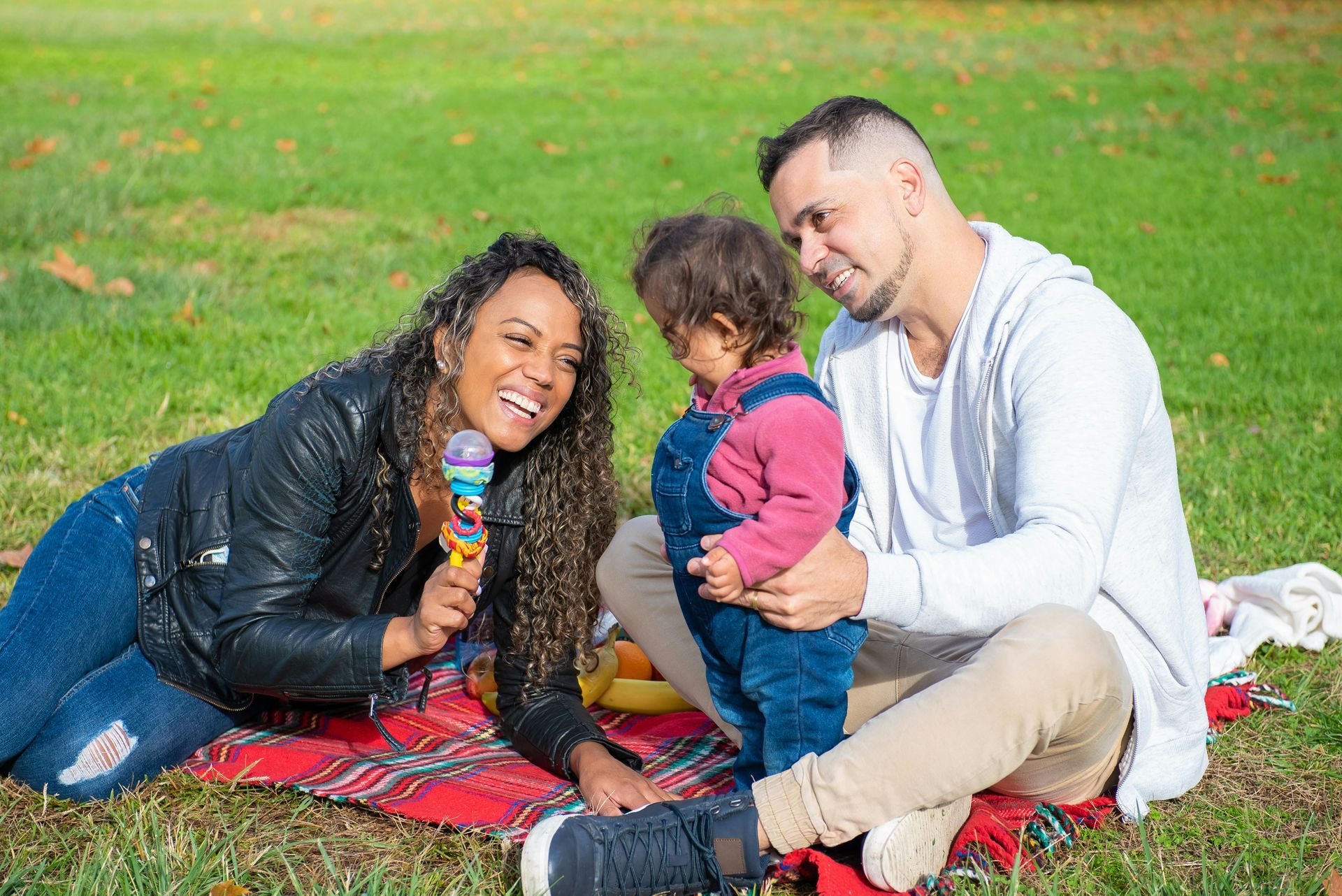 A family of three sitting on a red blanket in a grassy park, smiling while interacting with a toddler holding a toy.