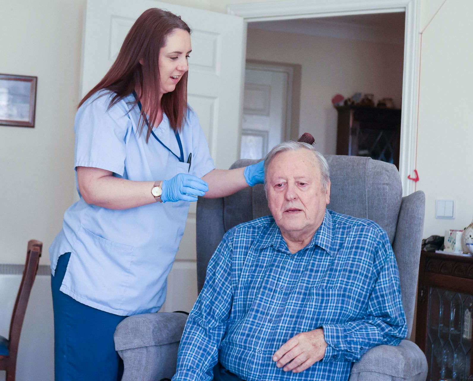 A healthcare worker in blue scrubs and gloves tends to a person seated in an armchair in a home setting.