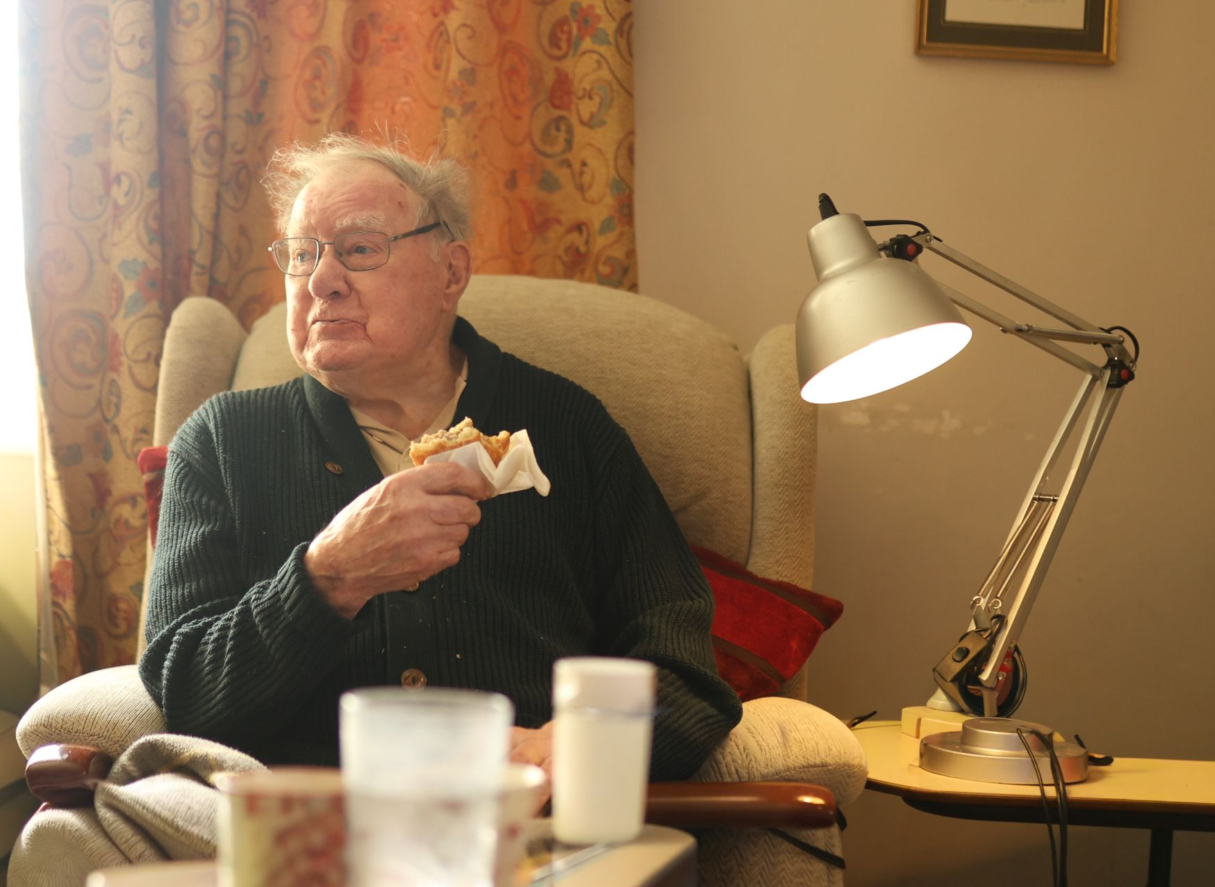 An older person wearing glasses eats a sandwich while seated in an armchair next to a desk lamp.