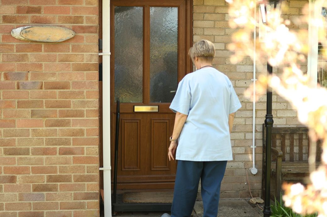A person in a light blue shirt and dark pants stands before a brown front door with a nameplate labeled “GRAZENA.”