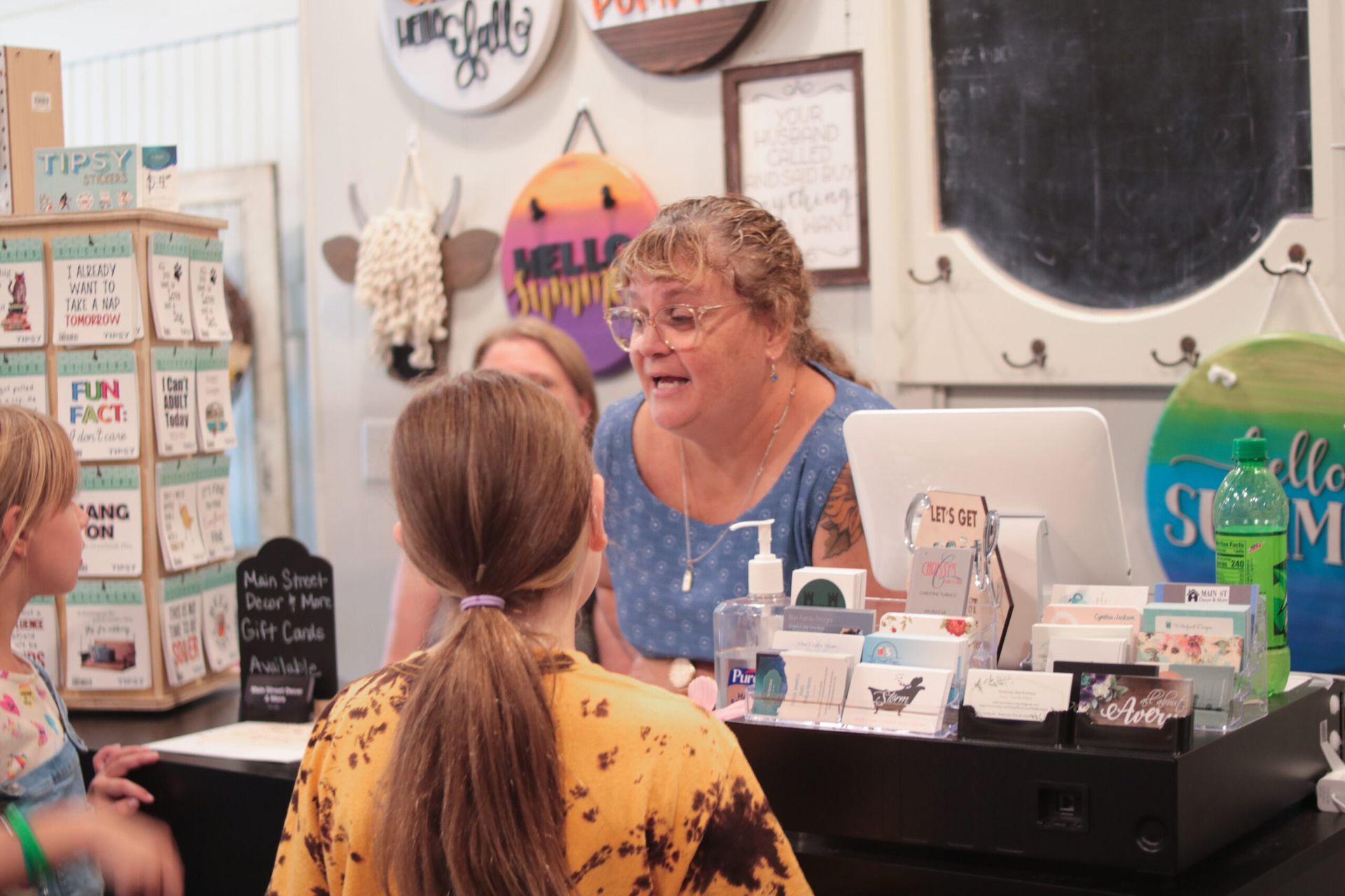 Woman speaking to customers at a shop counter, surrounded by merchandise.