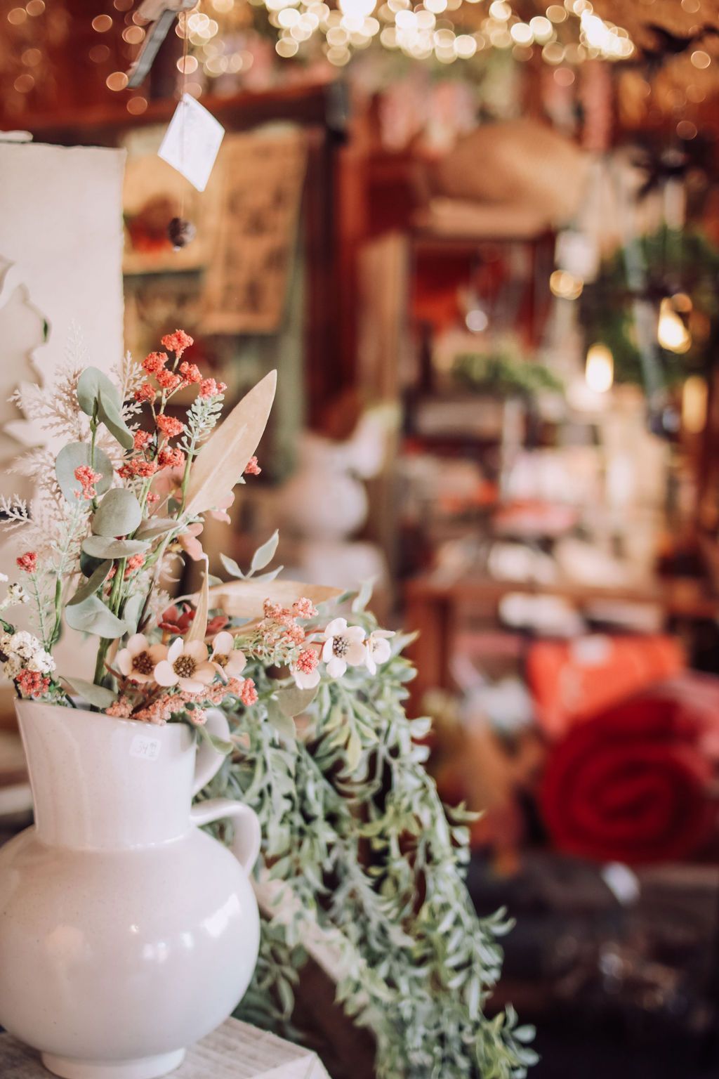 White vase with flowers, greenery in a shop with blurred merchandise and warm lighting.