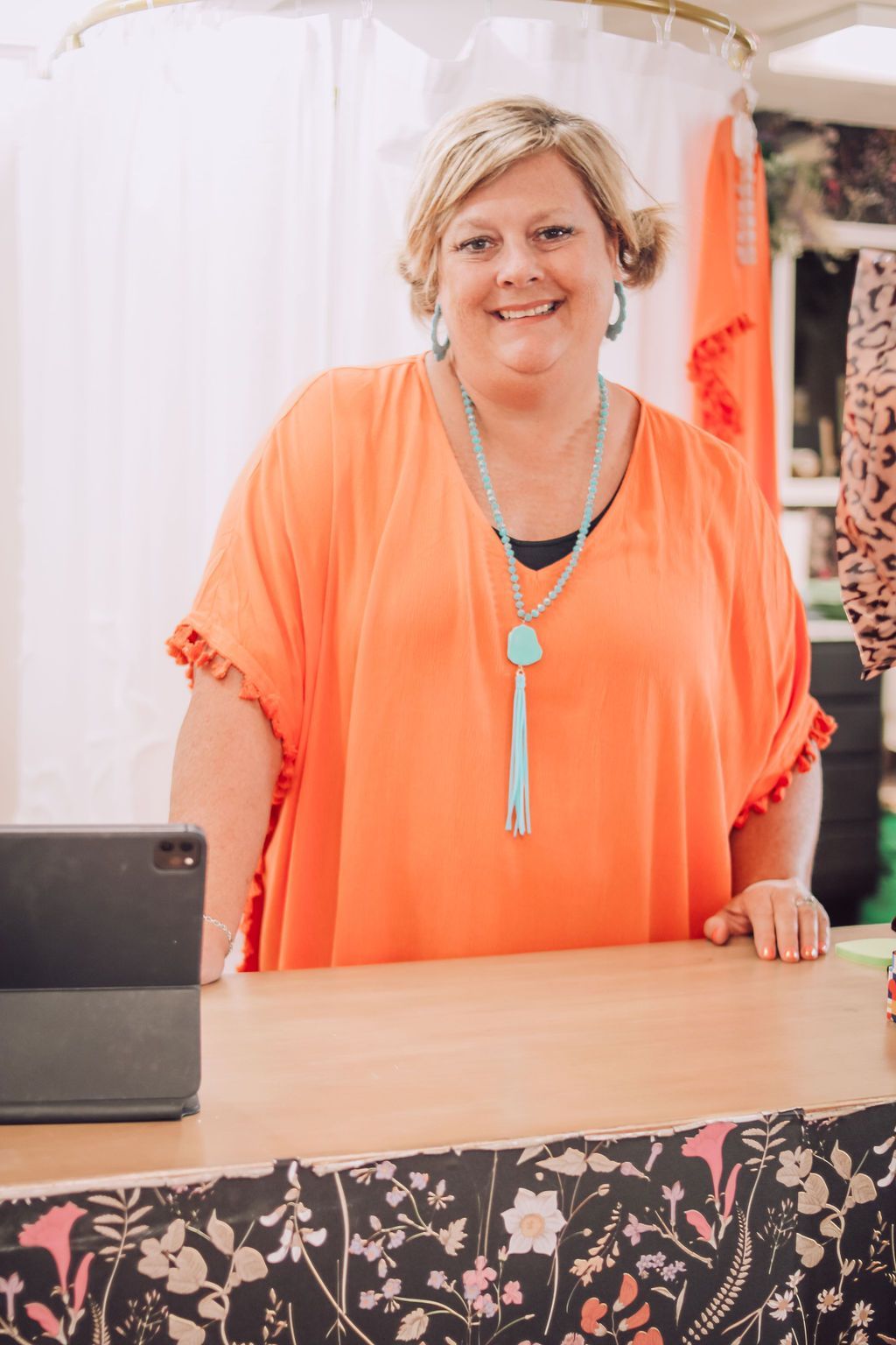 Woman in orange top smiling, turquoise necklace. Standing behind a counter with floral design.