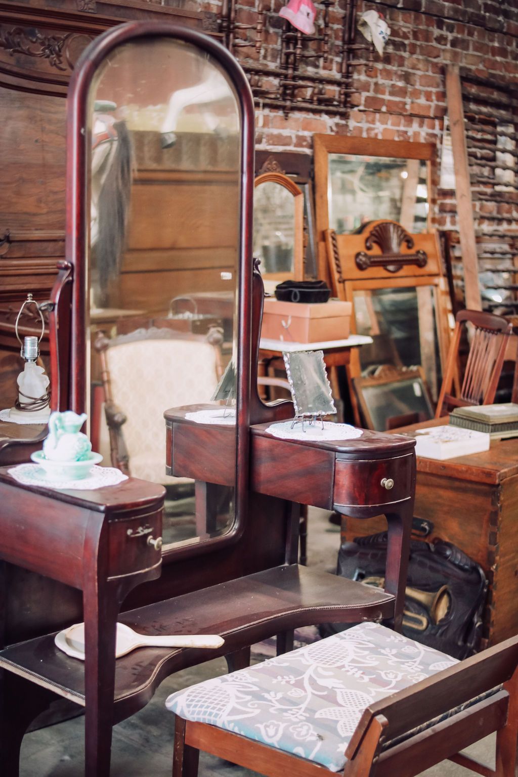Antique wooden vanity with tall mirror, drawers, and stool in a cluttered antique shop.