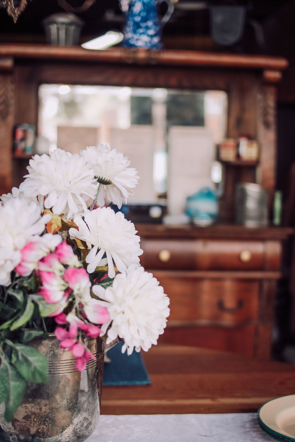 Bouquet of white and pink flowers in vase on table with blurred antique cabinet and mirror in background.