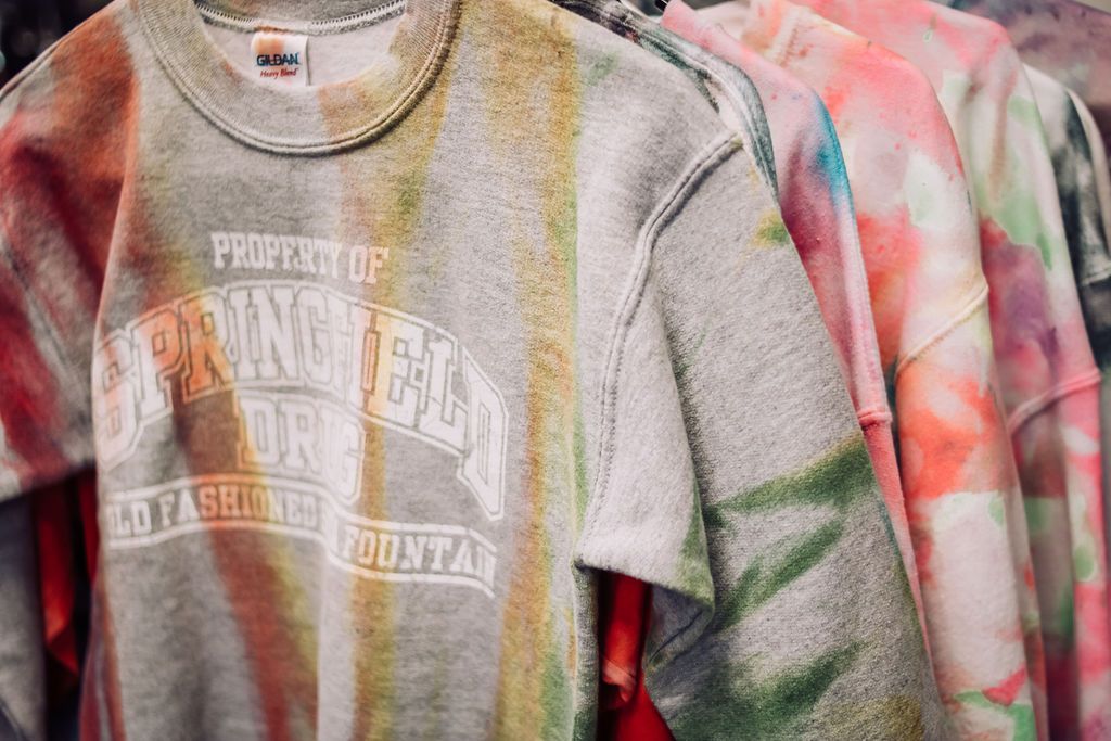 Tie-dye sweatshirts in various colors hanging on a rack; the first one is gray with faded lettering.