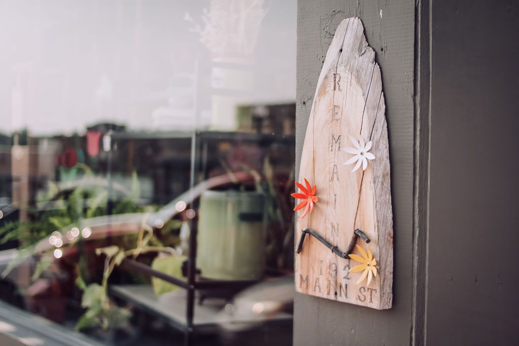 Wooden sign with flowers and text, mounted on a gray wall next to a window with plants visible inside.