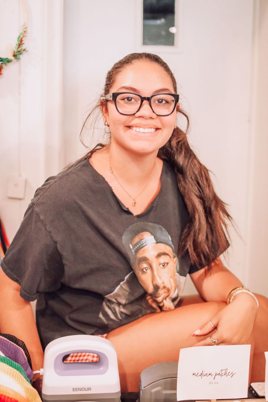 Woman wearing glasses and a Tupac shirt smiles, sitting near a crafting iron and a sign.