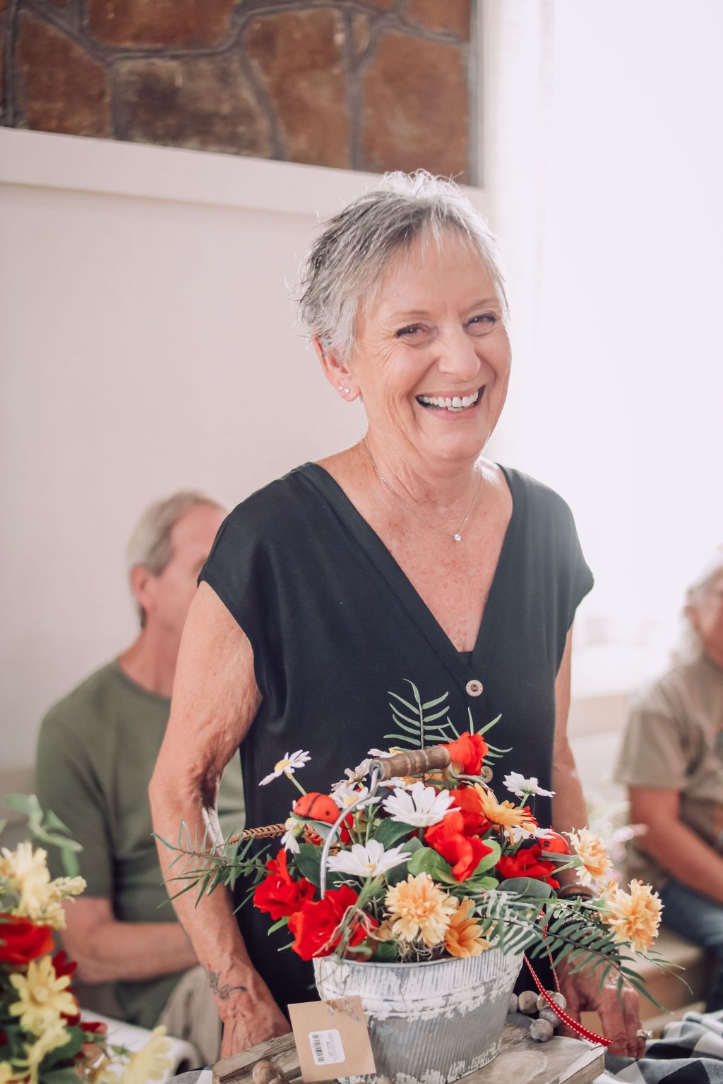 Woman smiling, holding flower arrangement. Black top, short gray hair, in bright room.