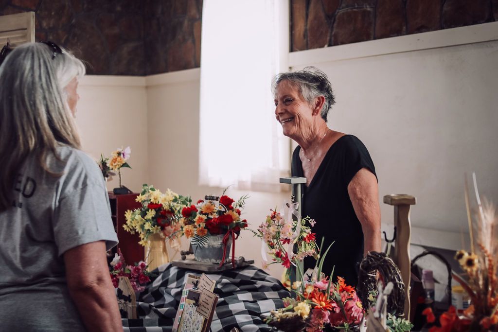 Two women near a table with flowers. One smiles, the other looks on. Indoors.