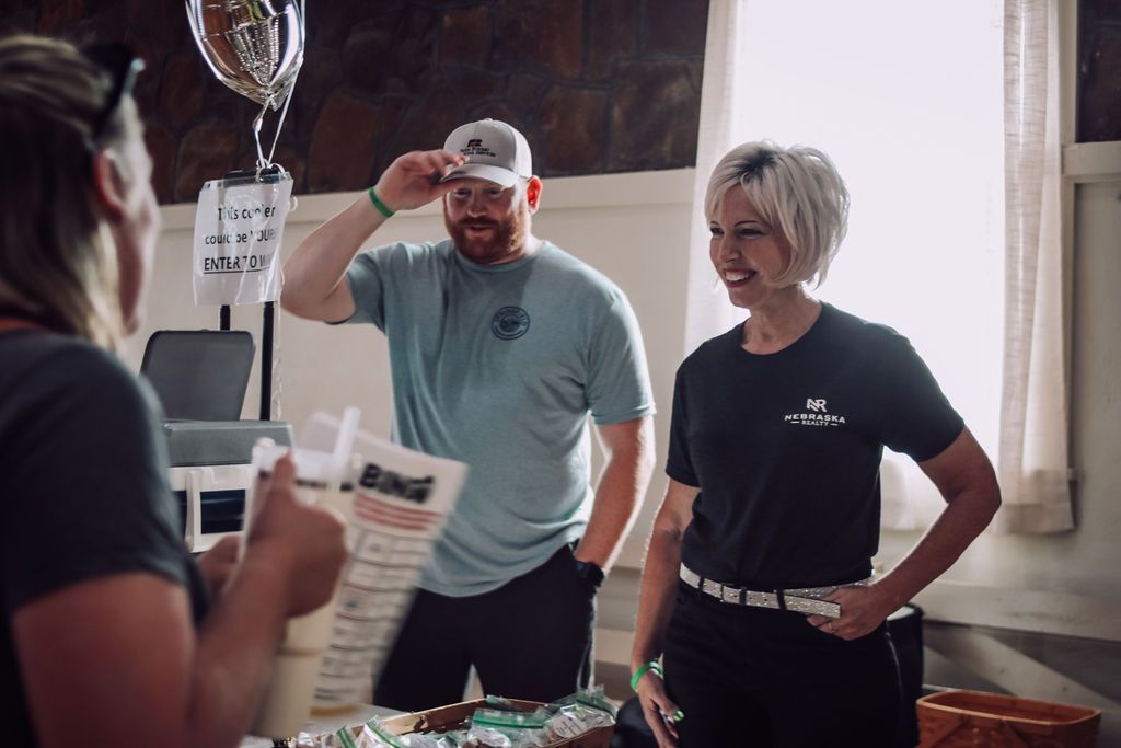 Three people at a table, one customer, two employees. Man in blue shirt holds a hat. Woman smiles. Indoors, natural light.