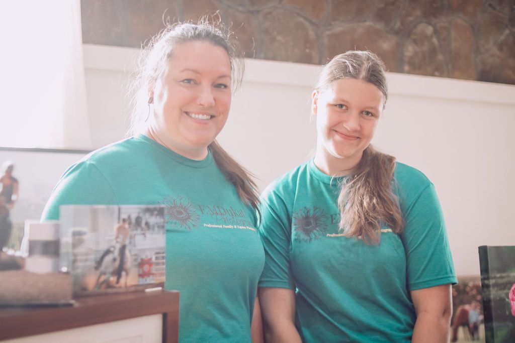 Two women wearing teal shirts smile, standing behind a table with photos in front of a neutral background.