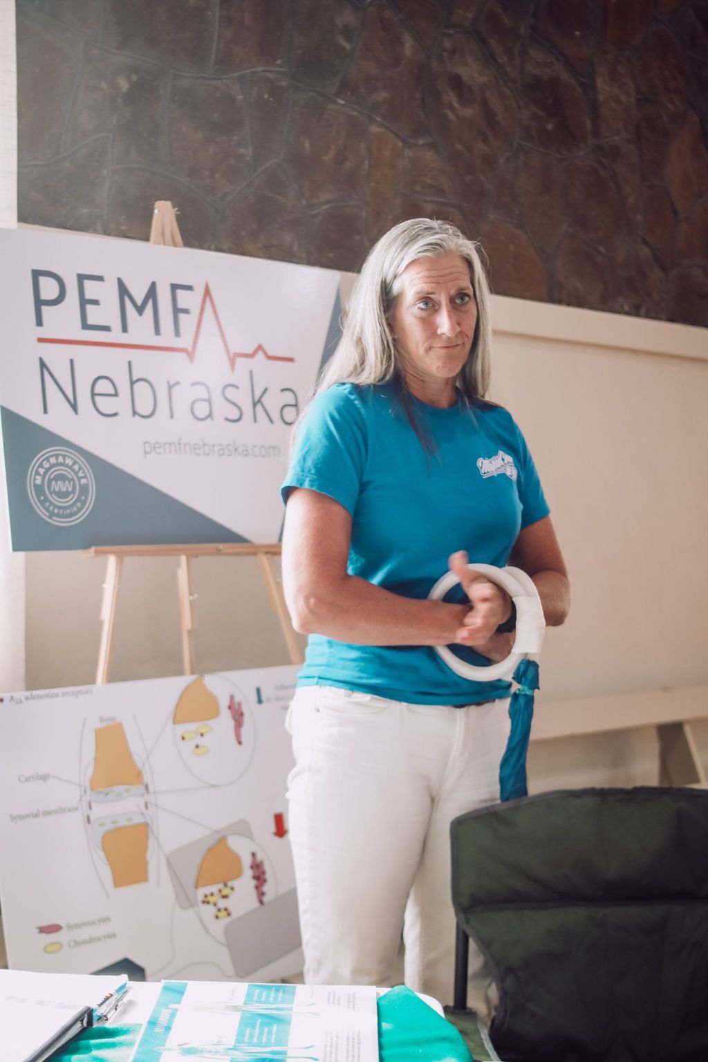 Woman in blue shirt, white pants, stands at a PEMF Nebraska display, holding a white device.