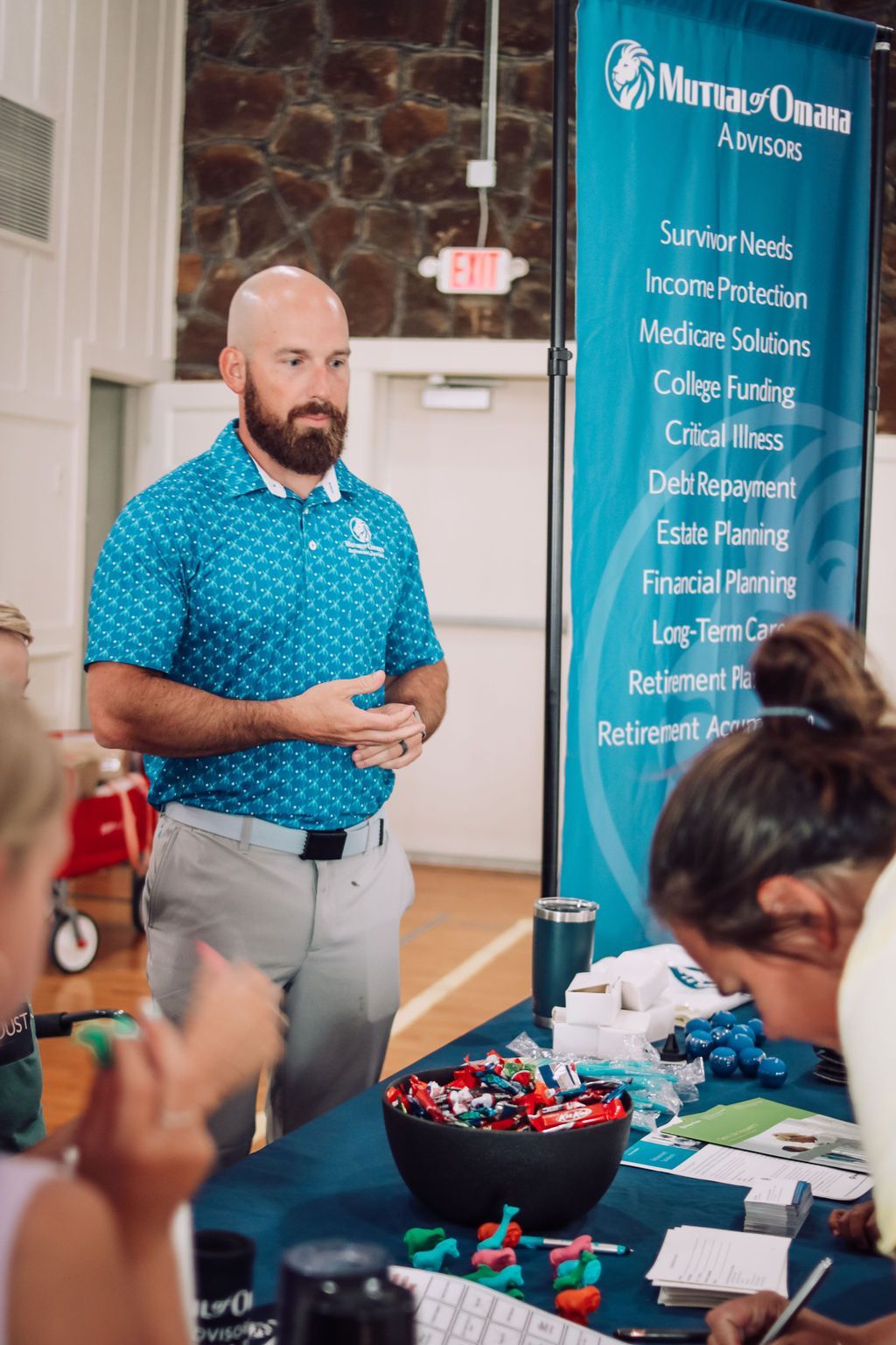 Man in blue shirt speaks at a table with a banner for financial services.