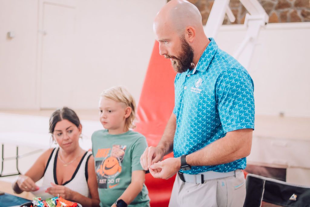 Man in blue shirt demonstrates to a boy and woman at a table; indoor setting.
