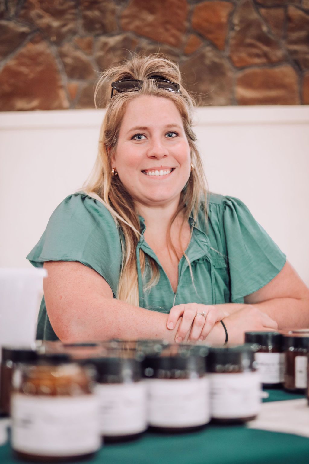Woman with blonde hair smiling behind jars of product at a market.