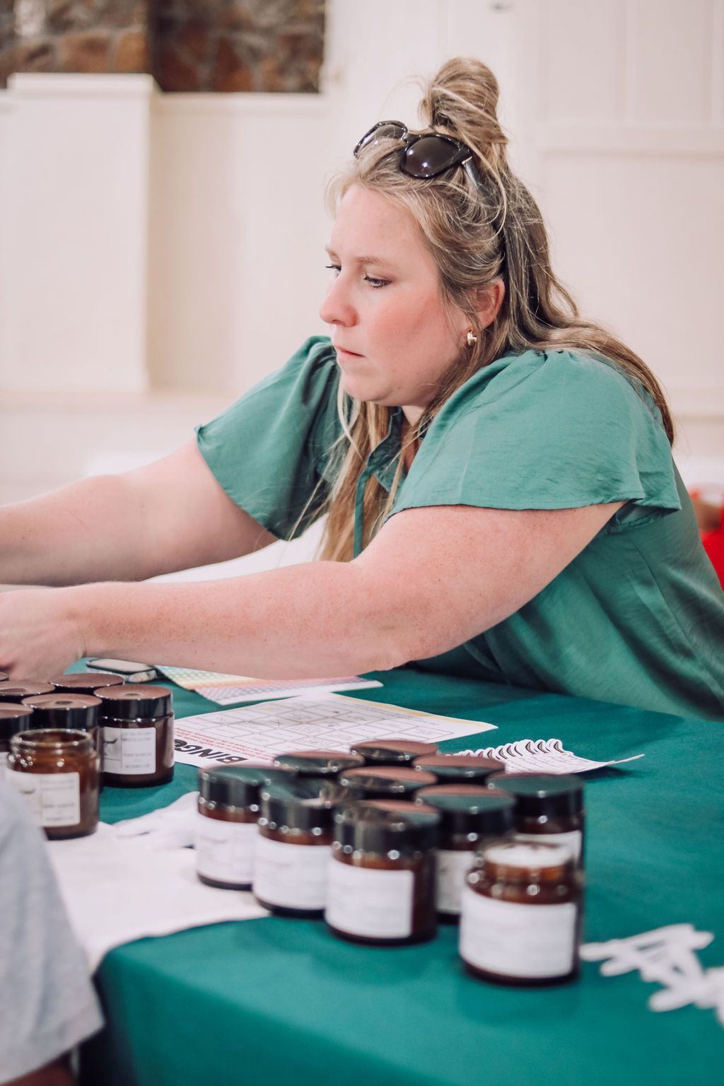 Woman arranging jars on a green table. She wears a green top, has sunglasses on her head, and is focused.