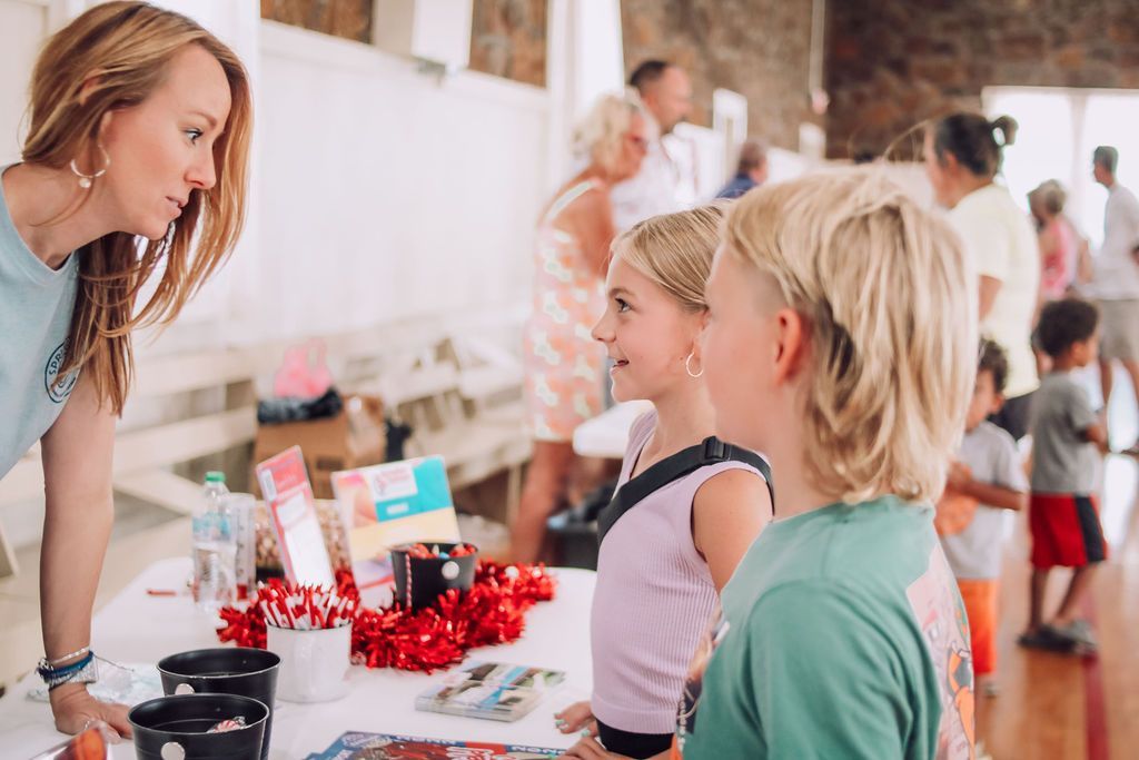 Woman speaks to two children at a table with items, others in background.